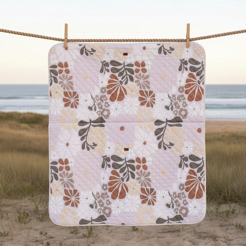 A rectangular picnic beach mat with a floral pattern in pastel pink, brown, and gray lies on the sand of a beach, with the ocean and sky in the background.