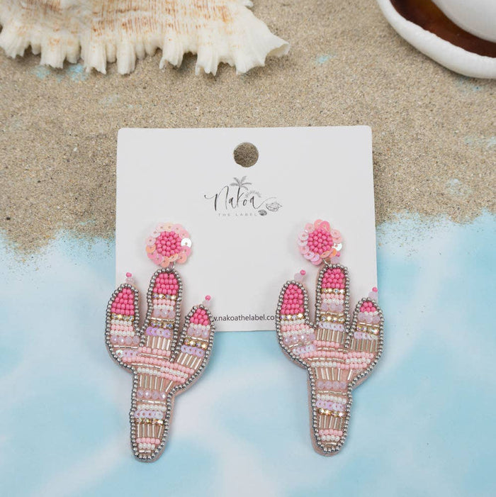 Pink and white beaded cactus earrings on a white display card, styled on a sandy surface with seashells.