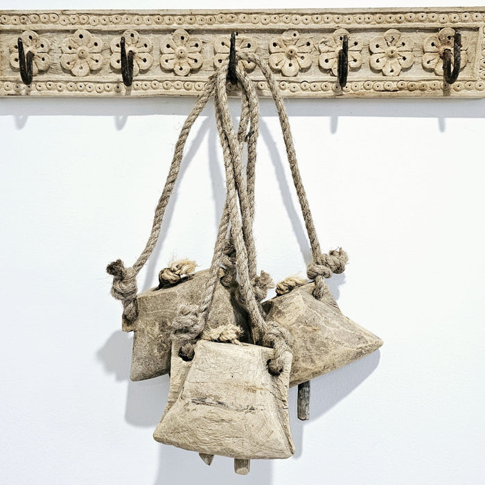 A vintage Indian wooden cow bell with a beige finish, featuring intricate designs, attached to a rope hanger. The bell and wood display signs of wear and age.