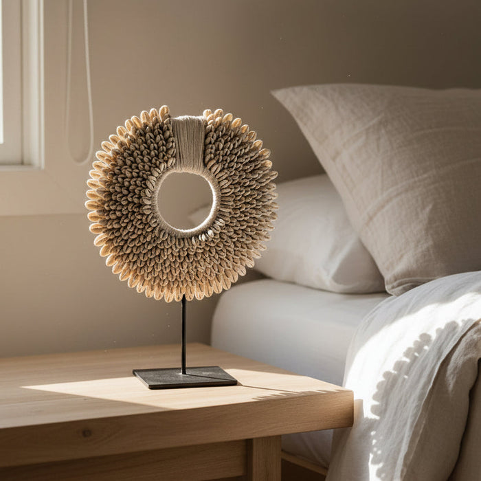 A light wood sideboard cabinet in a minimalist room, featuring a circular woven decorative sculpture on a black stand. White sheer curtains hang to the right, and light wood flooring is visible below.